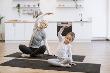 Yoga minderindeki sıska Müslüman kadın, küçük kız çocuğu ile çalışırken kollarını kapalı uzatıyor. Spor giyim sektöründe genç bir ebeveyn ve kız spor yaparken eğleniyor..