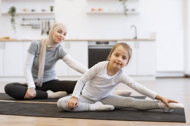 Yoga minderinde, kollarını ve bacaklarını esneten sıska müslüman kadın küçük kız çocukla antrenman yapıyor. Spor giyim sektöründe genç bir ebeveyn ve kız spor yaparken eğleniyor..