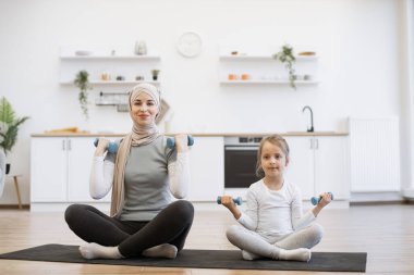 Duygusal müslüman kadın ve çocuk spor kıyafetleri içinde odada otururken heyecanla ağırlık kaldırıyorlar. Mutlu genç anne ve kız bebek evde neşe içinde egzersiz yapıyorlar..