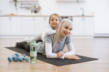 Egzersiz sırasında pozitif enerjiyle annelerin sırtüstü uzanan spor kıyafetli sevimli kız. Başörtülü müslüman bir kadın evde spor yaptıktan sonra tatlı bir kızla rahatlıyor..