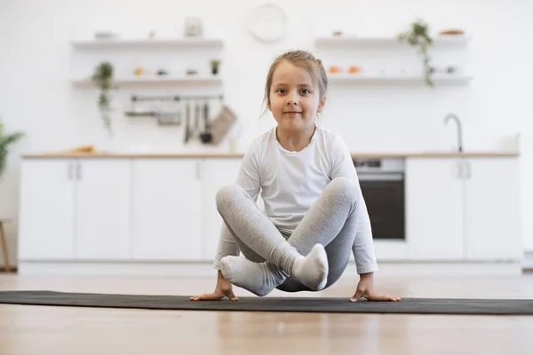 stock image Front view of cute girl practicing yoga, standing in crane exercise, bakasana pose, working out on mat wearing sportswear, indoor full length, in white loft kitchen background.