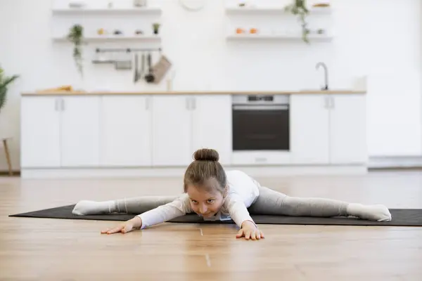 Caucasian preschool girl 5 years old in sports wear is engaged in gymnastics at home in the kitchen floor. Little cute female lying down on twine on mat in bright spacious room , copy space.