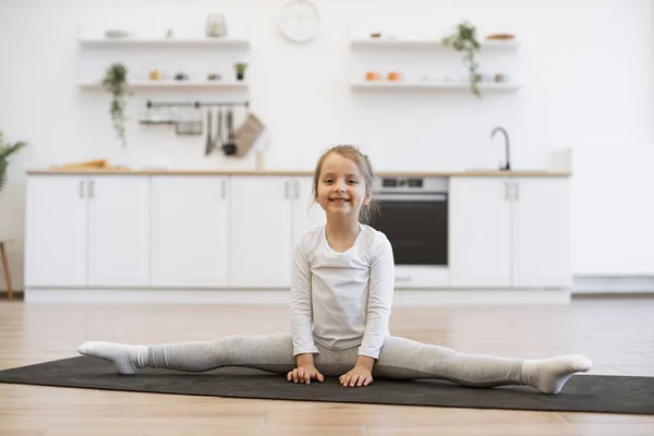 Beautiful caucasian girl 4 years old in a gymnastic wear is engaged in gymnastics at home in the kitchen floor. Little cute female sitting on twine on mat, copy space.