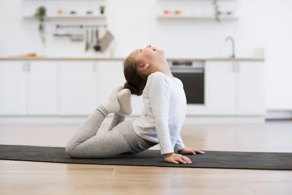 Sporty cute female sitting on mat and stretching by touching tips of legs to head in bright spacious room , copy space. Side view of preschool girl in sports wear is engaged in gymnastics at home.