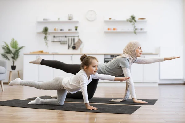 Active slim muslim lady and her daughter in sports clothes streching in balancing table pose on mats at home. Beautiful mother with her girl practicing yoga exercise together in kitchen.