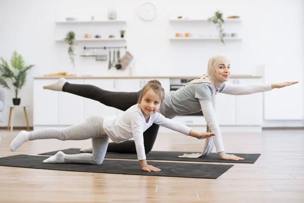Active slim muslim lady and her daughter in sports clothes streching in balancing table pose on mats at home. Mother with her girl practicing yoga exercise together in kitchen looking at camera.
