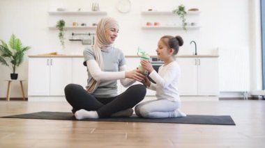 Aktif giyimli güzel Müslüman kadın elinde bir şişe su tutarken tatlı kızı lastik minderde otururken içki içiyor. Sağlıklı kadın ebeveyn ve küçük çocuk jimnastik sırasında sıvı kaybını telafi ediyorlar..