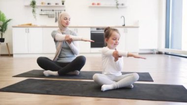 Yoga minderinde küçük bir kızın eğitim sırasında gözleri kapalı sıska müslüman bir bayanla kollarını açması ön planda. Spor giyim sektöründe genç bir ebeveyn ve kız spor yaparken eğleniyor..