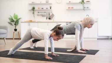 Çekici, tesettürlü Müslüman anne sabah uyandıktan sonra küçük kızıyla birlikte mutfakta tahta pozisyonu üzerinde çalışıyor. Çok rahat ve rahatım. Yoga yapan bir kadın ve küçük kızı sağlık hizmetleri için.