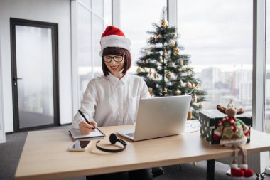 Noel Baba şapkalı genç bir iş kadını, elinde kalemle dizüstü bilgisayarın yanına yerleştirilen kâğıt taslağını not alıyor. Noel ağacının arka planındaki tahta masanın üzerine..