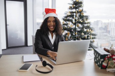Noel Baba şapkalı Afrikalı iş kadını Noel ağacının yanında oturan kameraya bakıyor ve tatil sezonunda dizüstü bilgisayarda çalışıyor, bilgisayar klavyesinde daktilo ediyor..