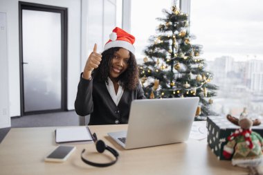 Noel Baba şapkalı Afrikalı iş kadını kameraya bakıyor, başparmağını kaldırıyor, Xmas ağacının yanındaki ofiste oturuyor ve tatil sezonu boyunca dizüstü bilgisayarda çalışıyor, klavyede daktilo yazıyor..