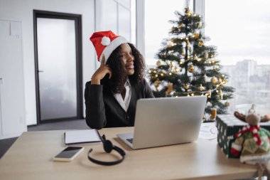 Noel Baba şapkalı düşünceli Afrikalı iş kadını Noel ağacının yanında oturuyor ve tatil sezonunda dizüstü bilgisayarda çalışıyor, bilgisayar klavyesinde daktilo kullanıyor..