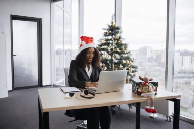 Noel Baba şapkalı Afrikalı bir iş kadını Noel ağacının yanında oturuyor ve tatil sezonunda dizüstü bilgisayarda çalışıyor, bilgisayar klavyesinde yazı yazıyor..