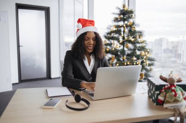 Noel Baba şapkalı Afrikalı bir iş kadını Noel ağacının yanında oturuyor ve tatil sezonunda dizüstü bilgisayarda çalışıyor, bilgisayar klavyesinde yazı yazıyor..