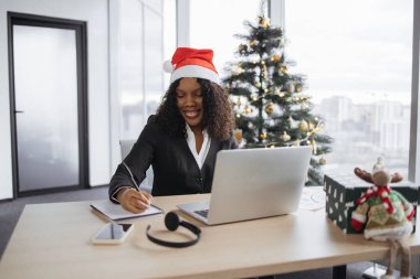 Noel Baba şapkalı genç Afrikalı iş kadını, elinde kalemle dizüstü bilgisayarın yanına yerleştirilen ve Noel ağacının arka planındaki tahta masanın üzerine not alıyor..
