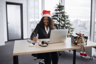 Noel Baba şapkalı genç Afrikalı iş kadını, elinde kalemle dizüstü bilgisayarın yanına yerleştirilen ve Noel ağacının arka planındaki tahta masanın üzerine not alıyor..