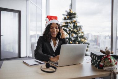 Noel ağacının arka planında taşınabilir bilgisayar kullanırken akıllı telefondan konuşan neşeli Afrikalı kadın. Verimli serbest çalışan kadın tatillerde uzak şirketlerin müşterisiyle konuşuyor..