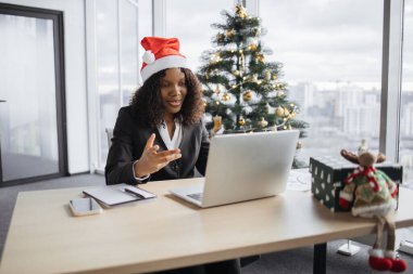 Takım elbiseli mutlu Afrikalı iş kadını Noel ağacının arka planında müşteriyle konuşuyor. Noel Baba şapkalı kendine güvenen yönetici internet uygulaması kullanarak projenin yeni detaylarını açıklıyor.