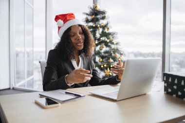 Takım elbiseli mutlu Afrikalı iş kadını Noel ağacının arka planında müşteriyle konuşuyor. Noel Baba şapkalı kendine güvenen yönetici internet uygulaması kullanarak projenin yeni detaylarını açıklıyor.