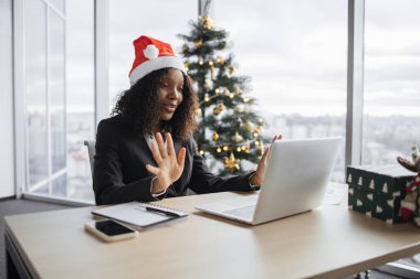 Takım elbiseli mutlu Afrikalı iş kadını Noel ağacının arka planında müşteriyle konuşuyor. Noel Baba şapkalı kendine güvenen yönetici internet uygulaması kullanarak projenin yeni detaylarını açıklıyor.