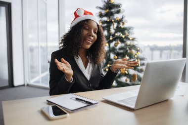 Takım elbiseli mutlu Afrikalı iş kadını Noel ağacının arka planında müşteriyle konuşuyor. Noel Baba şapkalı kendine güvenen yönetici internet uygulaması kullanarak projenin yeni detaylarını açıklıyor.
