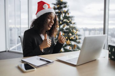 Takım elbiseli mutlu Afrikalı iş kadını, Noel ağacının arka planında başparmak göstererek müşteriyle görüntülü konuşma yapıyor. Noel Baba şapkalı işletmeci projenin yeni detaylarını açıklıyor