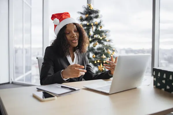 Happy African businesswoman in suit talking to client over video call on background of the Christmas tree. Confident business manager in Santa hat explaining new details of project using internet app.