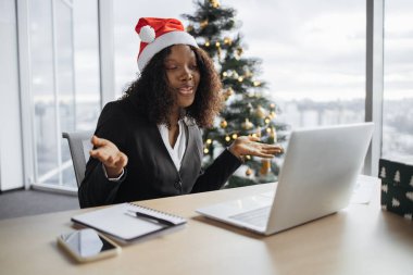 Takım elbiseli mutlu Afrikalı iş kadını Noel ağacının arka planında müşteriyle konuşuyor. Noel Baba şapkalı kendine güvenen yönetici internet uygulaması kullanarak projenin yeni detaylarını açıklıyor.