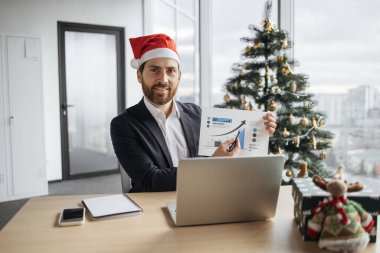 Kendine güvenen yetişkinler, Noel ağacının arka planında ofisten sunum yaparken, fotoğraf makinesine bakarak, grafik materyallerini işaret ediyorlar..