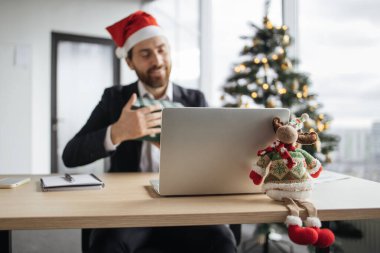 Masadaki Noel geyiğine odaklanın, Noel Baba şapkalı gülümseyen beyaz adamın bulanık arkaplanı çalışma masasında dizüstü bilgisayarla Noel ağacıyla süslenmiş bir şekilde otururken hediyesini açıyor..