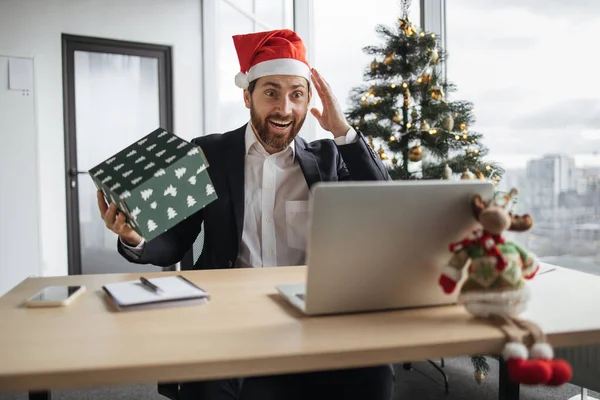 Excited Caucasian man in Santa hat unpacking Christmas gift while sitting at work desk with laptop. Happy male opening wrapped box and feeling excited during work office decorated with Christmas tree.