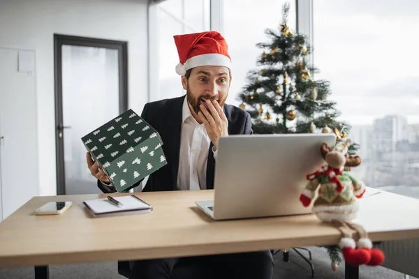 Excited Caucasian man in Santa hat unpacking Christmas gift while sitting at work desk with laptop. Happy male opening wrapped box and feeling excited during work office decorated with Christmas tree.