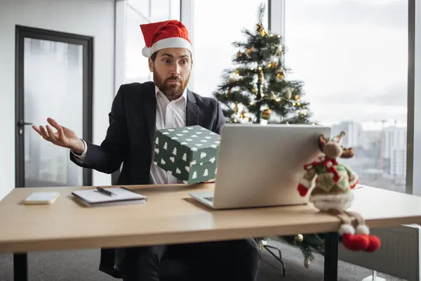 Caucasian business man in Santa hat holding with curiosity festive cardboard box in office decoraited Cristmas tree. Handsome male blogger making unpacking online order while recording video on laptop