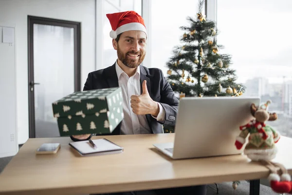 First person view of smiling bearded man in Santa hat and suit showing festive cardboard box with online order and thumb up while sitting at desk at office decorated with Christmas tree.