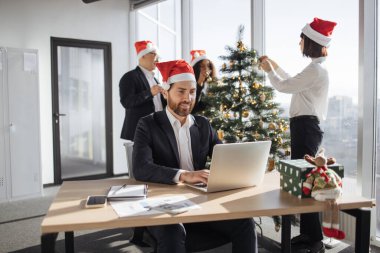 Takım elbiseli ve Noel Baba şapkalı çekici Kafkas işadamı Noel süslü ofiste dizüstü bilgisayar kullanarak masada oturuyor. İş yerindeki genç şirket çalışanı bilgisayar ekranına bakıyor..