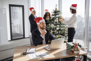 Takım elbiseli ve Noel Baba şapkalı çekici Kafkas işadamı Noel süslü ofiste dizüstü bilgisayar kullanarak masada oturuyor. İş yerindeki genç şirket çalışanı bilgisayar ekranına bakıyor..