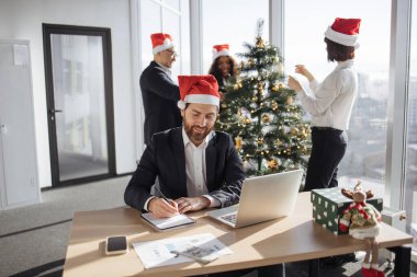 Noel Baba şapkalı genç işadamı, iş arkadaşları Noel ağacını süslerken dizüstü bilgisayarın yanına yerleştirilen kağıt döküman taslağını not alıyor.