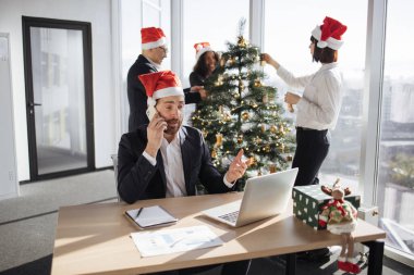 İş arkadaşlarının arka planında taşınabilir bilgisayar kullanırken akıllı telefondan konuşan neşeli beyaz işadamı Noel ağacını süslüyor.