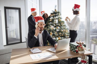 Takım elbiseli beyaz işadamı ve Noel Baba şapkası akıllı telefonla konuşurken taşınabilir dizüstü bilgisayar kullanarak Noel ağacını süsleyen meslektaşlarının arka planına bakıyor..