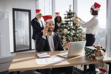 İş arkadaşlarının arka planında taşınabilir bilgisayar kullanarak akıllı telefondan konuşan neşeli beyaz işadamı Noel ağacını süslüyor..