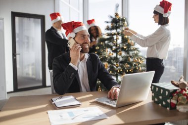 İş arkadaşlarının arka planında taşınabilir bilgisayar kullanarak akıllı telefondan konuşan neşeli beyaz işadamı Noel ağacını süslüyor..