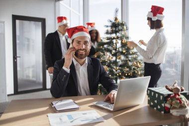 Beyaz takım elbiseli ve Noel Baba şapkalı bir işadamı, taşınabilir dizüstü bilgisayarla Noel ağacı süslemelerinin arka planında kameraya bakarken akıllı telefondan konuşuyor..