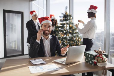 Beyaz takım elbiseli ve Noel Baba şapkalı bir işadamı, taşınabilir dizüstü bilgisayarla Noel ağacı süslemelerinin arka planında kameraya bakarken akıllı telefondan konuşuyor..