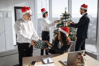 Asyalı ve Afrikalı Amerikalı meslektaşlar kış tatilini kutlamak için bayram ofisinde Noel ağacıyla Noel hediyesi kutusunu değiş tokuş ediyorlar. İnsanlar mevsimlik hediyeler veriyor ve Noel Baba hakkında neşeli hissediyorlar..