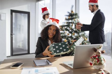 Güzel Afrikalı Amerikalı iş kadını Noel ağacını süsleyen iş arkadaşlarının arka planına bakarken masada oturup Noel hediyesini açıyor. .