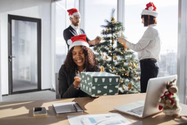 Takım elbiseli Afrikalı iş kadını portresi ve Noel Baba şapkası süpriz hediyeyi sunuyor. Süslü Noel ağacı ofisinde iş arkadaşlarıyla masada oturuyor..