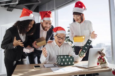 Gizli Noel Baba. Noel Baba şapkalı gülümseyen Asyalı işadamı, yanında duran çok uluslu meslektaşlarından aldığı bir Noel hediyesini açıyor. Yeni Yıl Arifesi.