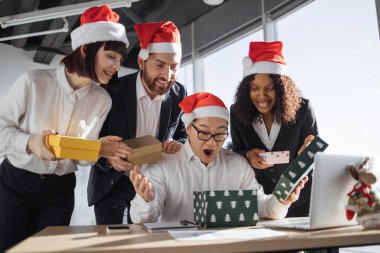 Secret Santa. Smiling Asian businessman in Santa hat unpacks a Christmas present from multinationals colleagues at work standing around him. New Years eve. clipart