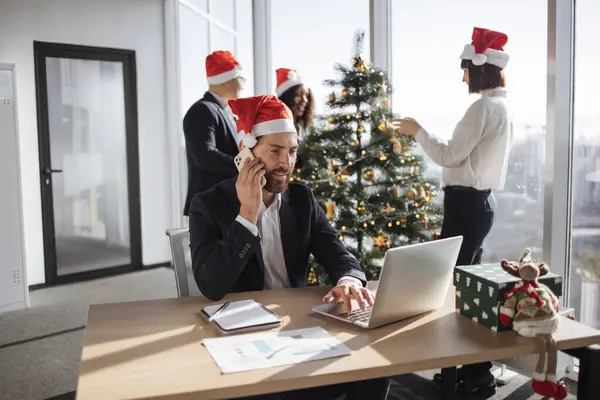 İş arkadaşlarının arka planında taşınabilir bilgisayar kullanırken akıllı telefondan konuşan neşeli beyaz işadamı Noel ağacını süslüyor.